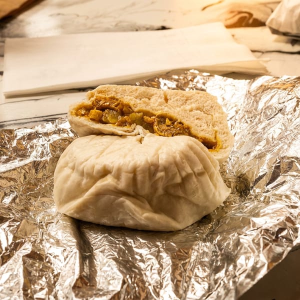 Two steamed buns, one cut open to reveal a savory filling, on crumpled aluminum foil.