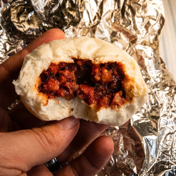 A hand holds a partially eaten steamed bun, revealing a savory red filling, over crinkled foil.