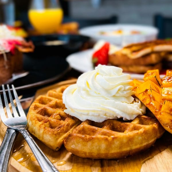 Golden waffle with whipped cream and sliced mango on a wooden plate, with other breakfast items blurred in the background.