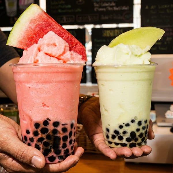 Hands holding two bubble tea smoothies, one pink with watermelon, one green with apple slices.