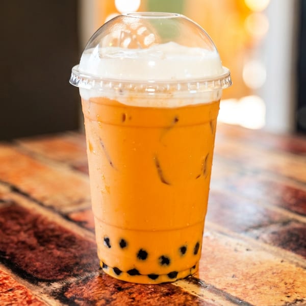 Iced Thai tea with boba pearls and a foamy top in a clear plastic cup on a brick-patterned table.