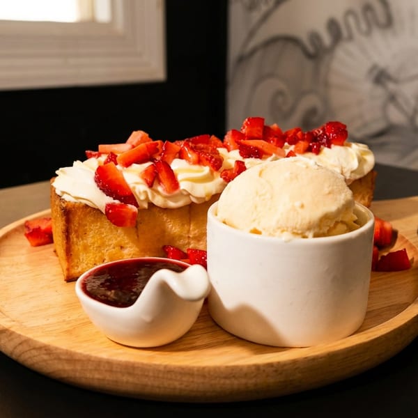 Golden toast with whipped cream, strawberries, vanilla ice cream, and berry jam on a wooden board.
